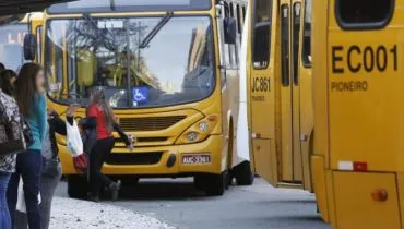 Na imagem, ônibus do sistema de transporte coletivo de Curitiba e pessoas aguardando no ponto para embarcar.