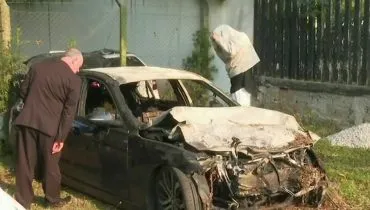 Imagem mostra uma BMW com a frente batida e queimada em um pátio da polícia. Duas pessoas estão em volta olhando.