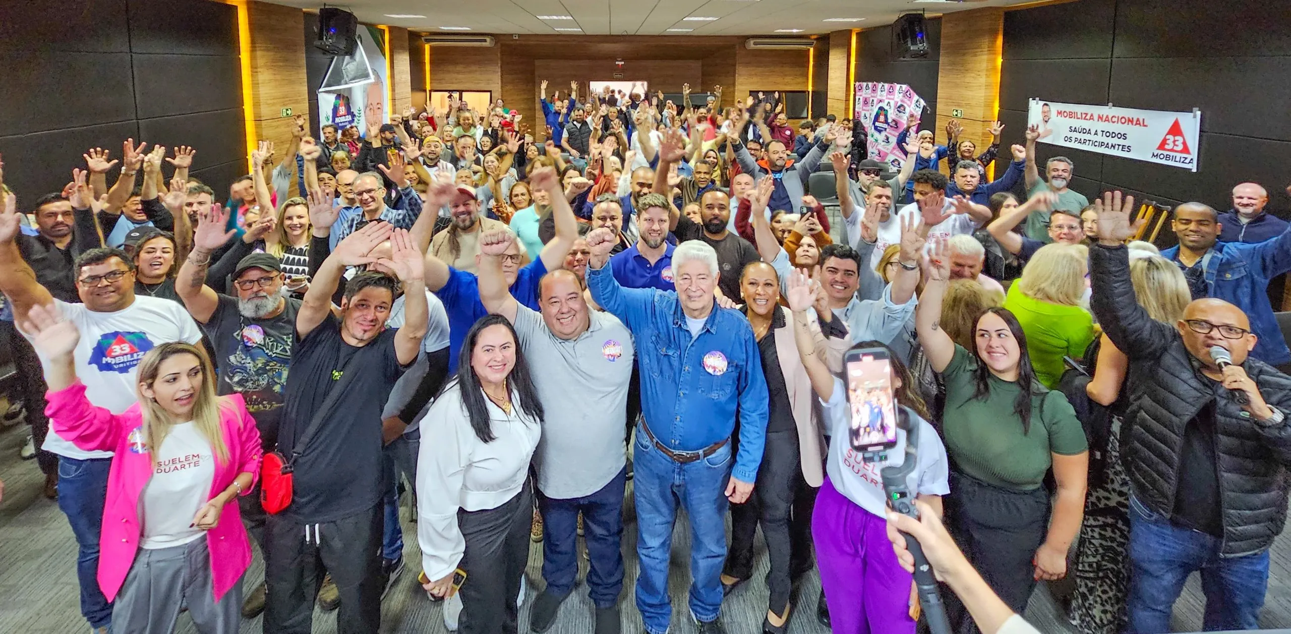 Imagem mostra quase uma centenas de pessoas no salão onde foi realizada a convenção. No centro, Requião de braça direito erguido posando para a foto com correligionários ao redor