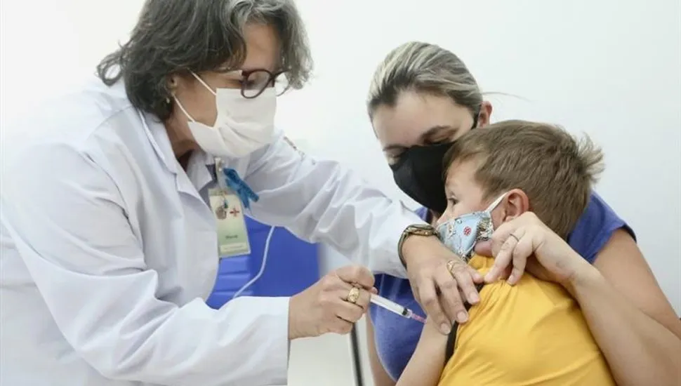 Profissional da saúde aplica injeção em criança, que está no colo da mãe.