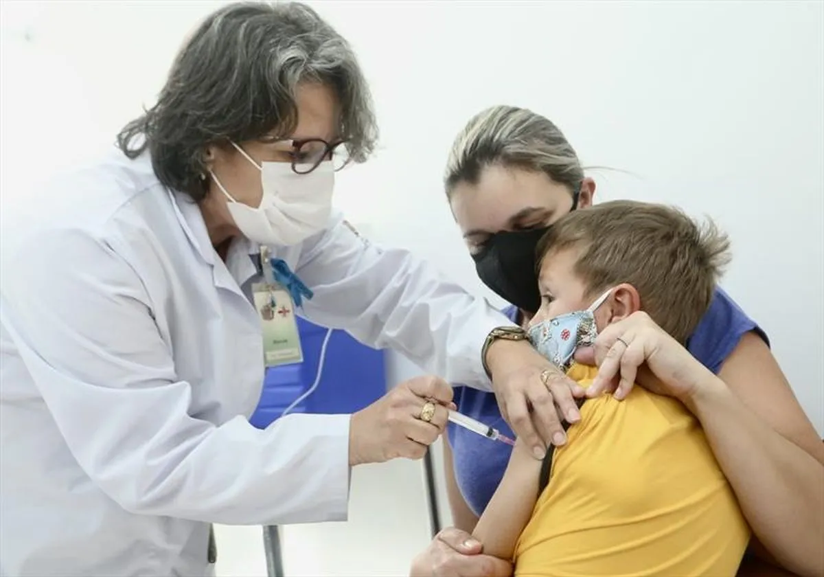 Profissional da saúde aplica injeção em criança, que está no colo da mãe.