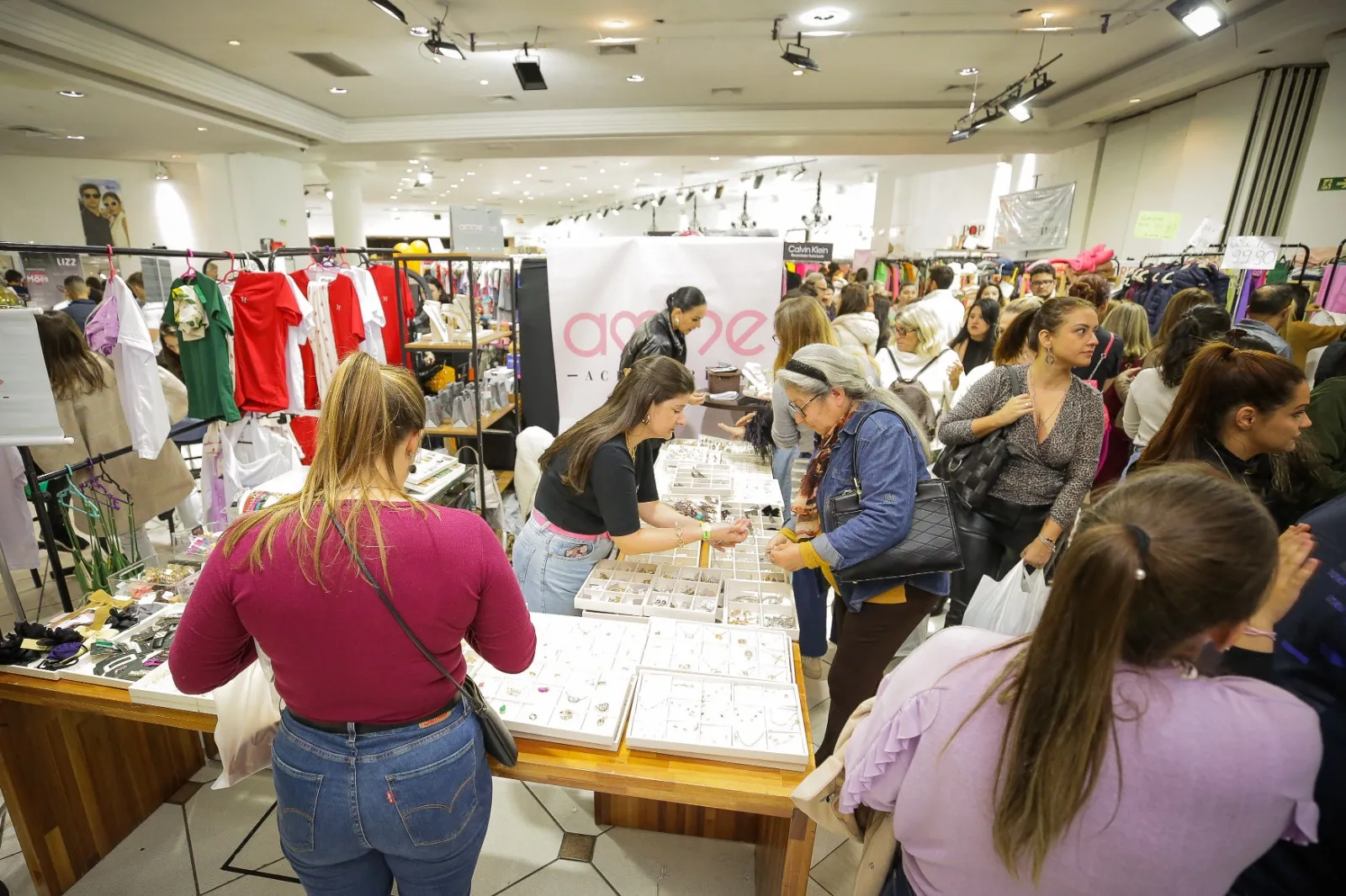 Foto mostra várias pessoas dentro de uma sala grande, com araras de roupas e mesas cheias de produtos