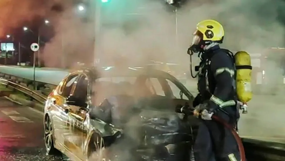 Imagem mostra um carro pegando fogo, com muita fumaça e um bombeiro uniformizado apagando o incêndio.