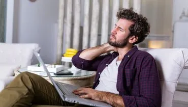 Foto mostra homem sentado num sofá, com um notebook no colo, reclamando de dores no pescoço