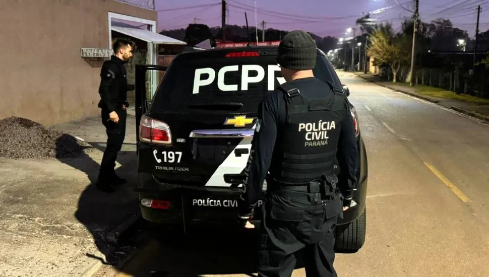 Imagem mostra uma viatura da Polícia Civil do Paraná durante a operação em uma rua, a noite, em Piraquara. Dois policiais estão de costas.