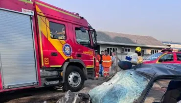 Imagem mostra parcialmente um carro que ficou destruído. Ao fundo populares olham a cena ao lado de um caminhão de bombeiros