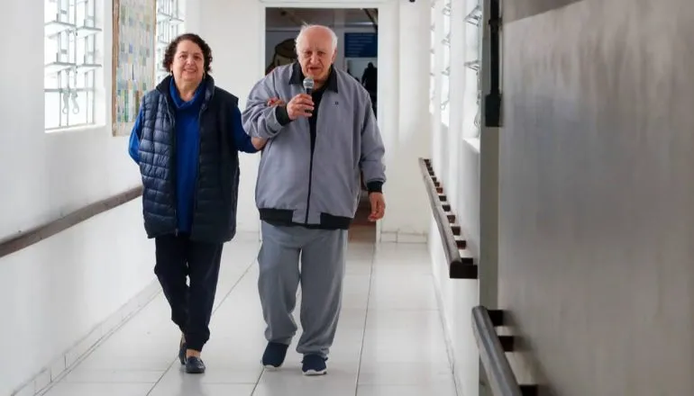 Imagem mostra um homem e uma mulher andando em um corredor. Ele está com um microfone na mão e agasalho cinza. Ela está de calça preta e blusa azul.