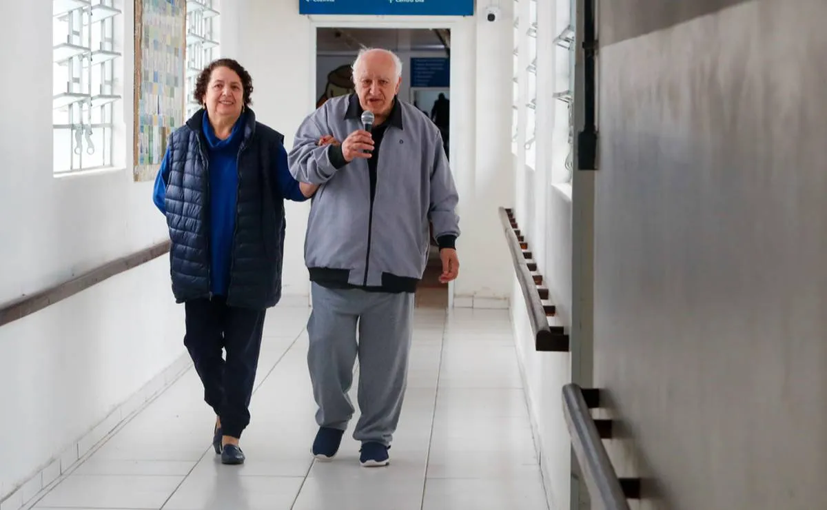 Imagem mostra um homem e uma mulher andando em um corredor. Ele está com um microfone na mão e agasalho cinza. Ela está de calça preta e blusa azul.