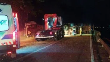 Imagem a noite mostra um caminhão tombado em uma rodovia, uma ambulância do lado esquerdo e luzes de farol.