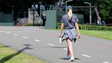 Imagem mostra uma mulher andando no Parque Barigui. Está muito calor e ela usa boné, bermuida e camiseta cinza. Ela está com uma garrafa de água na mão.