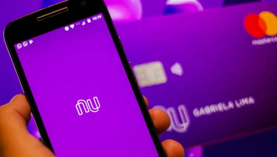 Na imagem, celular com aplicativo do Nubank aberto. Ao fundo, cartão do banco Nubank. Cor roxa predomina na foto.