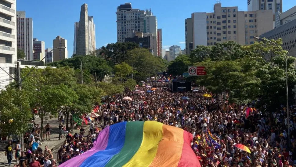 Imagem da Av. Cândido de Abreu, no Centro de Curitiba, com multidão e uma gigante bandeira o orgulho LGBT em cima de parte das pessoas.