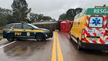 Caminhão tombando sobre pistas da rodovia, carro da PRF e ambulância na imagem.