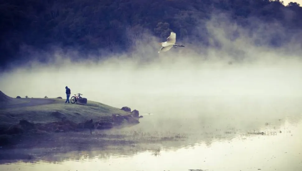 Imagem mostra um lago com vapor com um barranco com geada. Tem uma pessoa de bicicleta e uma ave voando bem no meio da foto.