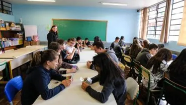 Imagem mostra uma sala de aula com alunos e um quadro verde ao fundo.