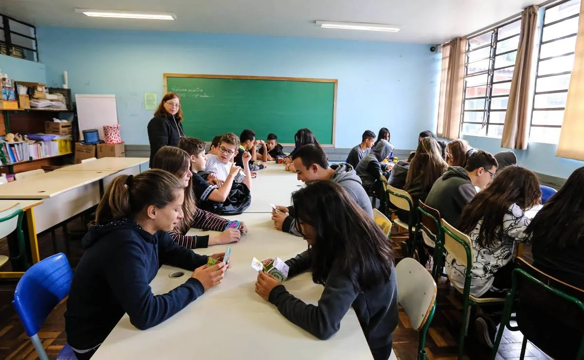 Imagem mostra uma sala de aula com alunos e um quadro verde ao fundo.