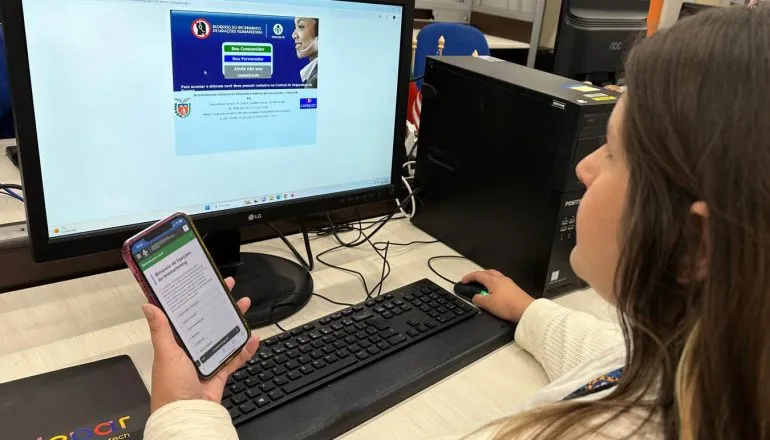 Mulher, em frente ao computador, com celular na mão.