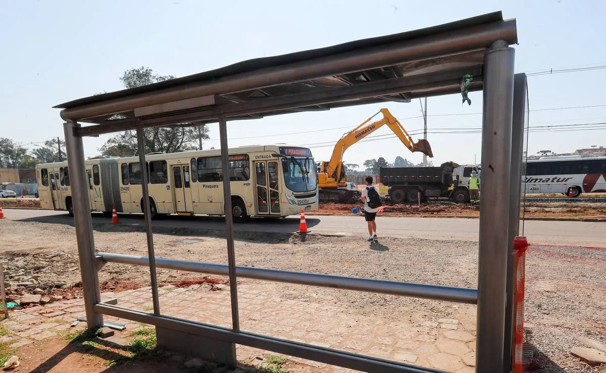 Imagem mostra um ponto de ônibus desmontado com um ônibus branco ao longe e uma máquina no fundo da imagem.