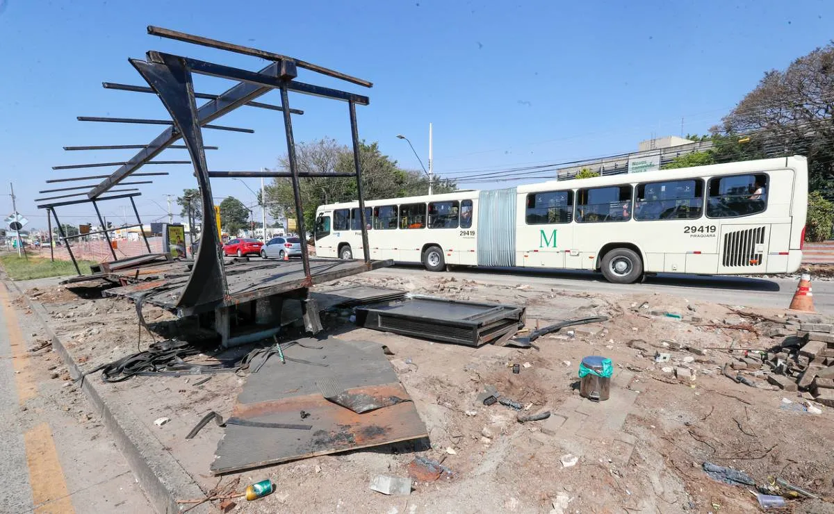 Imagem mostra um onibus branco passando ao lado de uma estação tudo desmontada com céu azul.