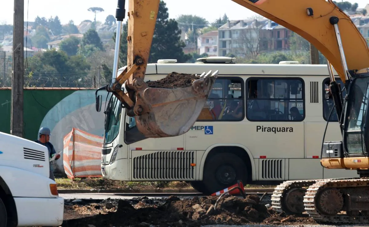 Imagem mostra uma pa carregadeira em primeiro plano com um ônibus branco ao fundo.