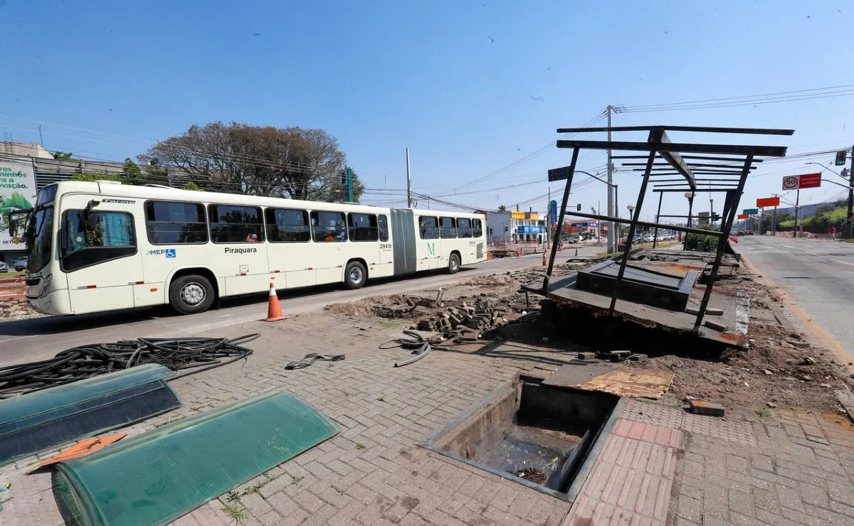 Imagem mostra um onibus branco passando ao lado de uma estação tudo desmontada com céu azul.