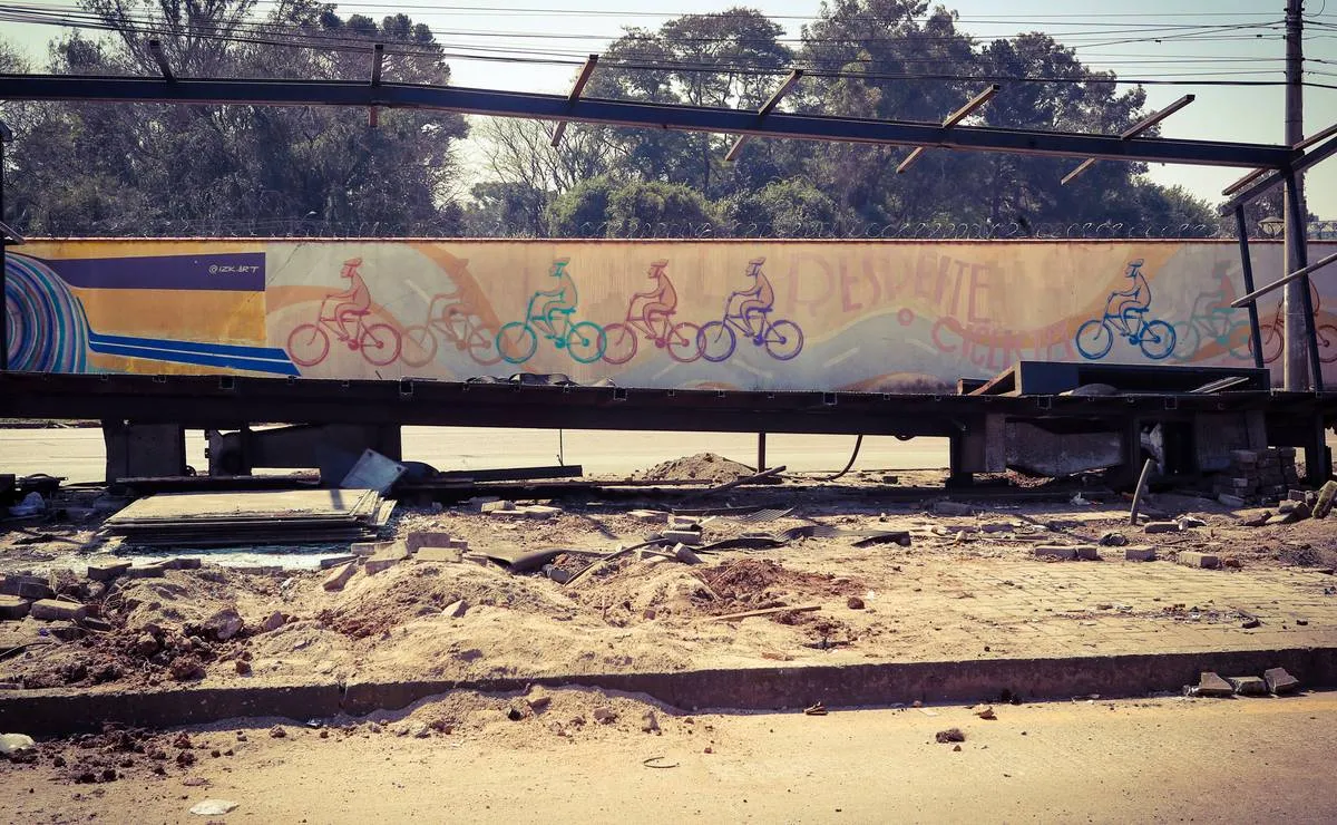 Imagem mostra uma estação tubo desmontada na frente de um muro com pintura de pessoas andando de bicicleta.