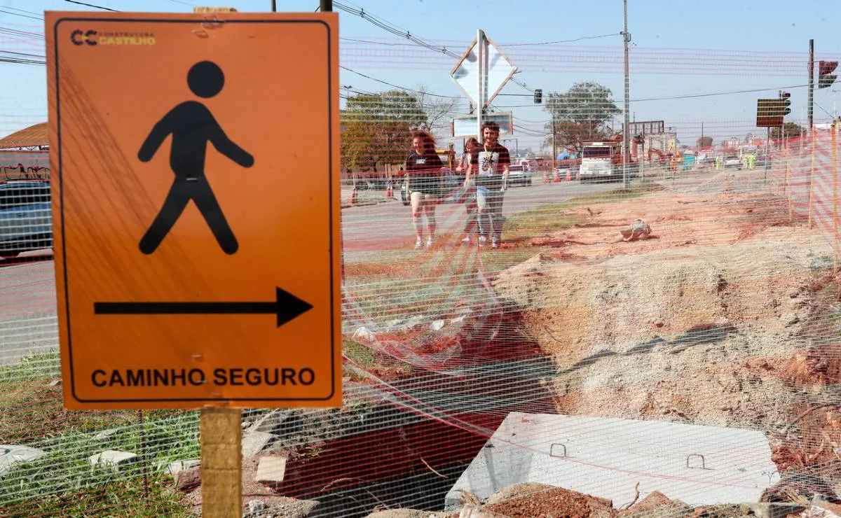 Imagem mostra uma placa alaranjada com o escrito "caminho seguro" para pedestres no meio de uma grande obra. 