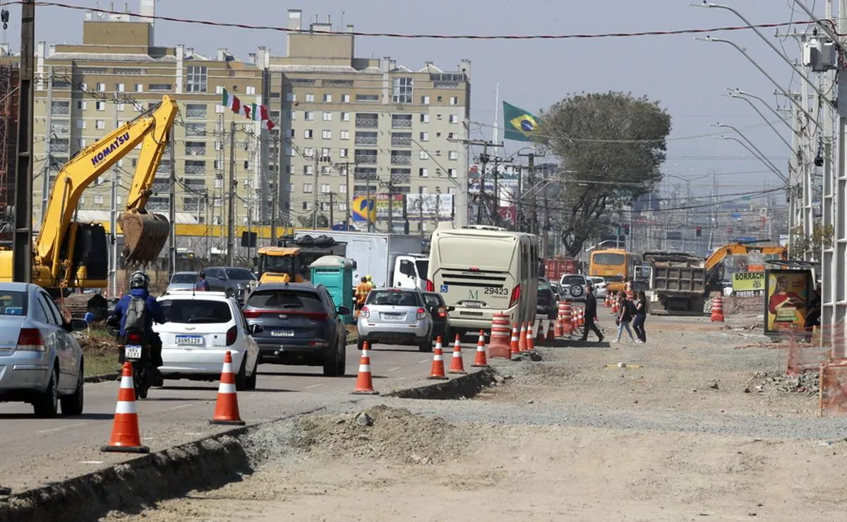 Imagem mostra o trânsito em uma avenida em obra em Curitiba com pedestres tentando pegar o ônibus.