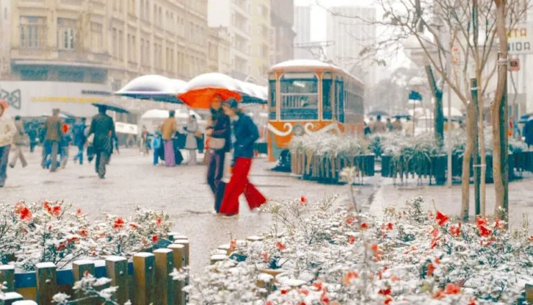 Imagem antiga mostra pessoas caminhando com neve em Curitiba, ao lado do Bondinho da Rua XV. As pessoas estão com muito frio e com guarda chuva para se proteger da neve.