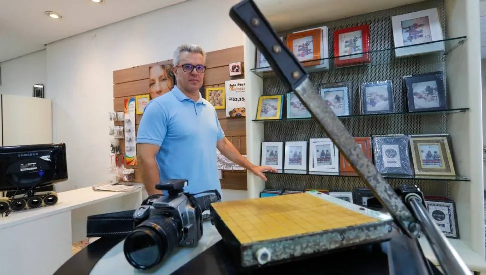 A imagem mostra um homem de camiseta azul olhando para o fotógrafo. A imagem tem uma guilhotina de cortar papel e uma câmera. Há porta retratos ao fundo.