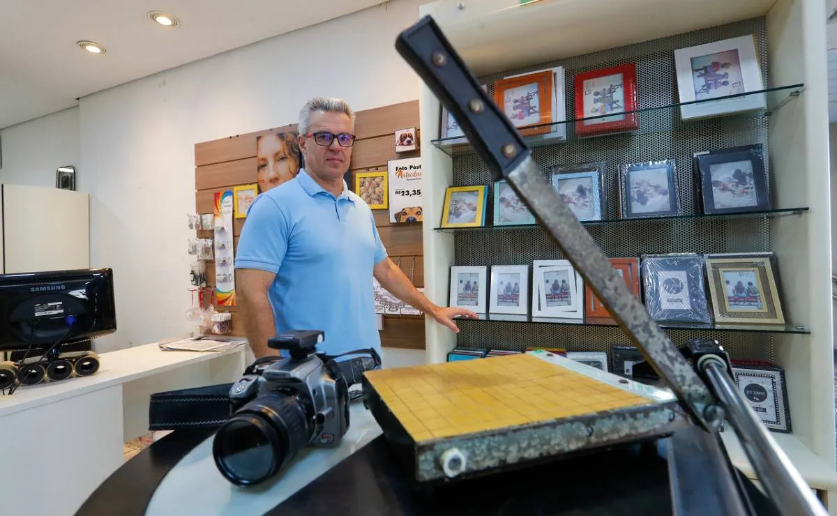 A imagem mostra um homem de camiseta azul olhando para o fotógrafo. A imagem tem uma guilhotina de cortar papel e uma câmera. Há porta retratos ao fundo.