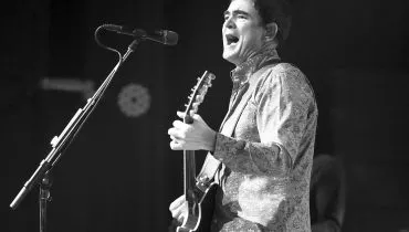 Foto mostra cantor samuel rosa tocando uma guitarra e cantando em um microfone num pedestal