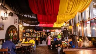 Interior do Bar do Alemão, em Curitiba. Mesas de madeira, lugares cheios, uma grande bandeira nas cores da Alemanha - vermelho, amarelo e preto - como decoração na parte superior, junto ao teto.