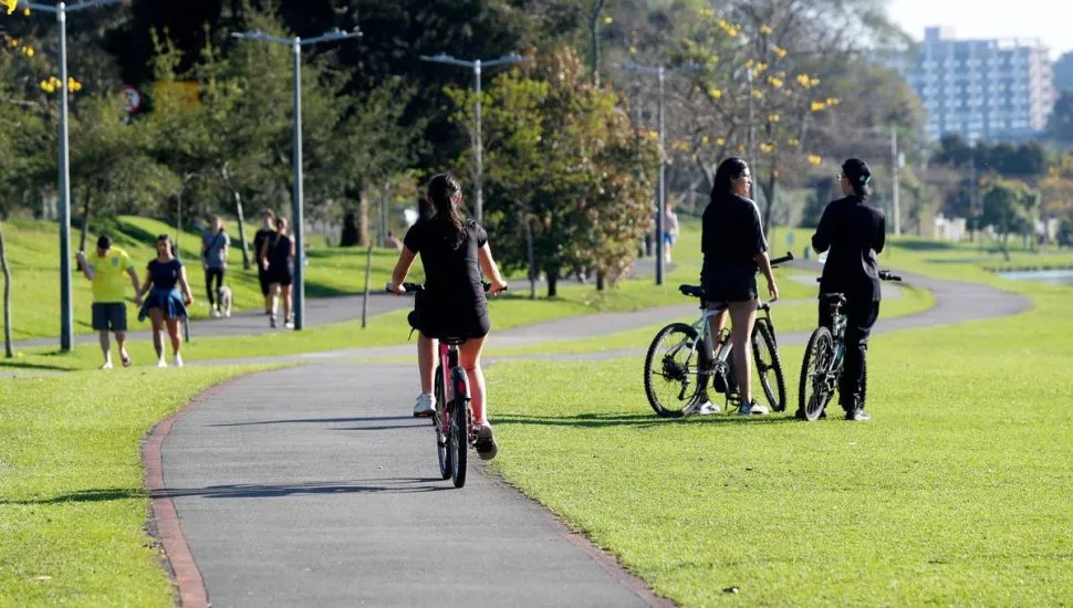 Imagem mostra um dia de sol no Parque Barigui, em Curitiba, com pessoas andando de bicicleta e conversando.