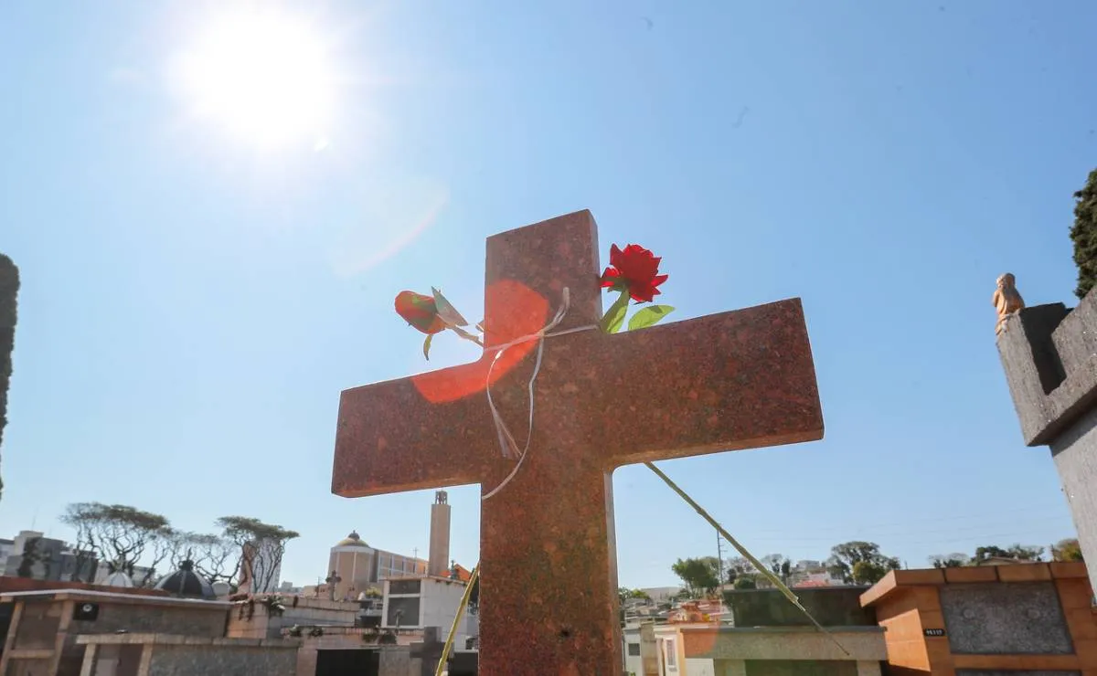 Imagem mostra uma cruz com céu azul ao fundo. Tem duas rosas de plástico amarradas nela.