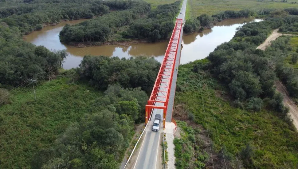 Na imagem, veículos passam em ponte metálica laranja, sobre um rio.