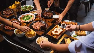 Imagem mostra três pessoas (só os seus braços) pegando comida numa mesa repleta de pratos com carnes assadas, batatas fritas e outros acompanhamentos