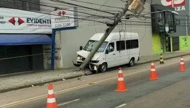 Imagem mostra uma van branca batida contra um poste. Há cones na pista e lojas fechadas.