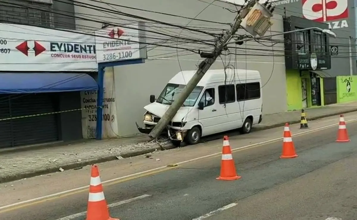 Imagem mostra uma van branca batida contra um poste. Há cones na pista e lojas fechadas.