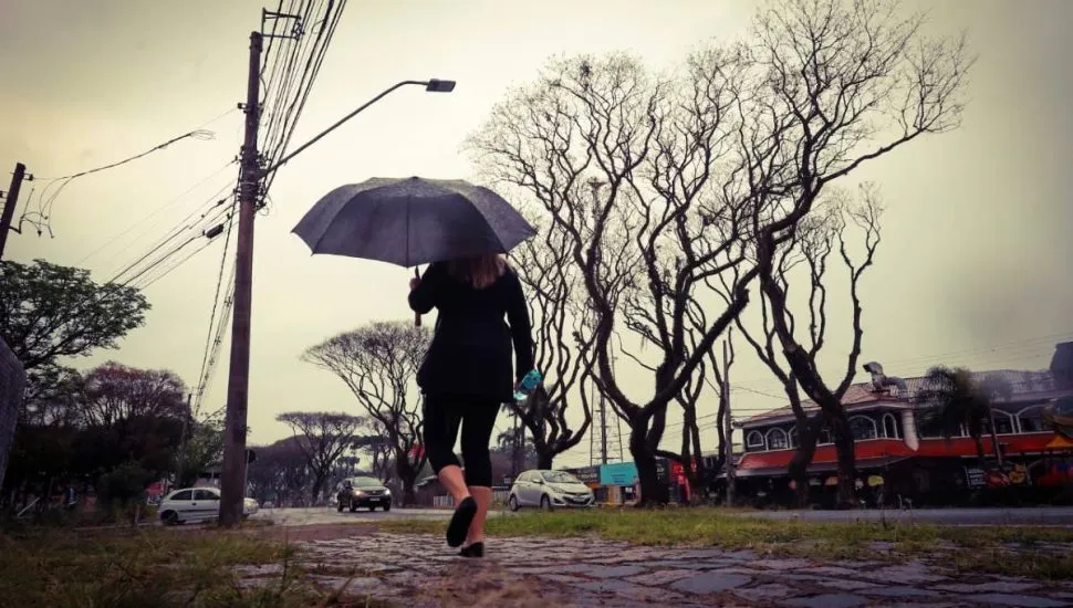 Imagem mostra uma mulher andando em uma calçada de Curitiba com um guarda-chuva. O tempo está fechado, o céu cinza e tem árvores secas no canteiro central.