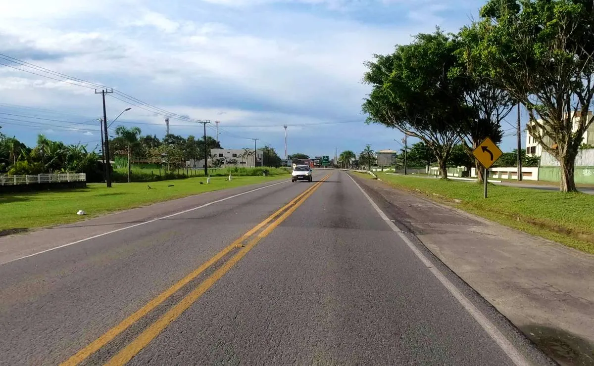 Imagem mostra uma estrada de pista simples com um carro e céu com nuvens.