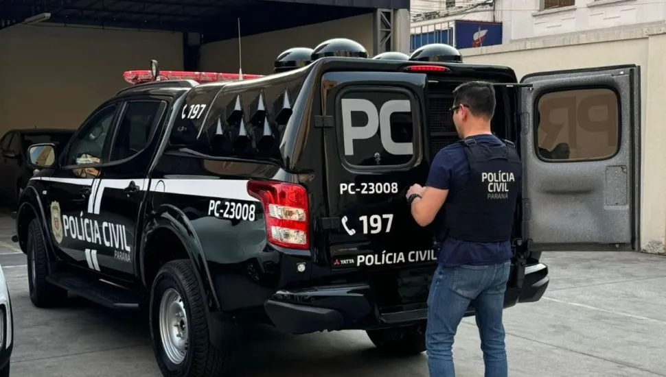 Na imagem, policial aparece em frente à porta traseira de um carro da polícia.