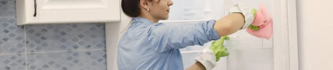 Imagem mostra uma mulher limpando uma geladeira. Ela usa luvas verdes e veste uma blusa azul.