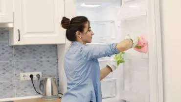 Imagem mostra uma mulher limpando uma geladeira. Ela usa luvas verdes e veste uma blusa azul.