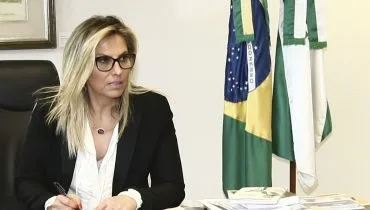 Na imagem, Cida Borghetti, sentada ao lado de bandeiras do Brasil e do Paraná.