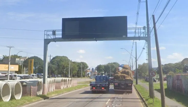 Imagem mostra um painel de trânsito em uma avenida. Tem dois caminhões passando por baixo e céu azul.