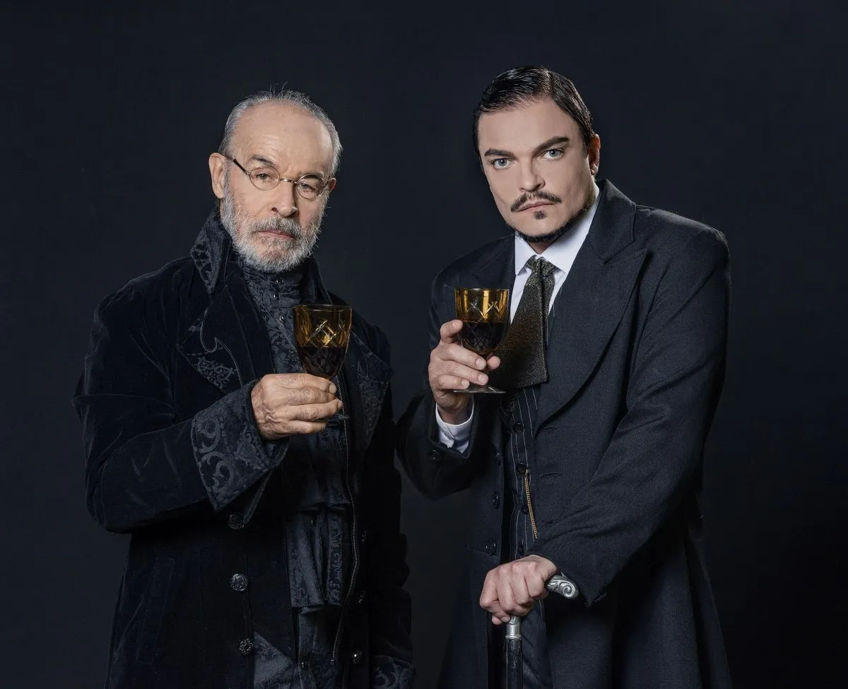 Foto mostra os atores Osmar Prato e Maurício Cardoso segurando uma taça de bebida na frente de um pano preto