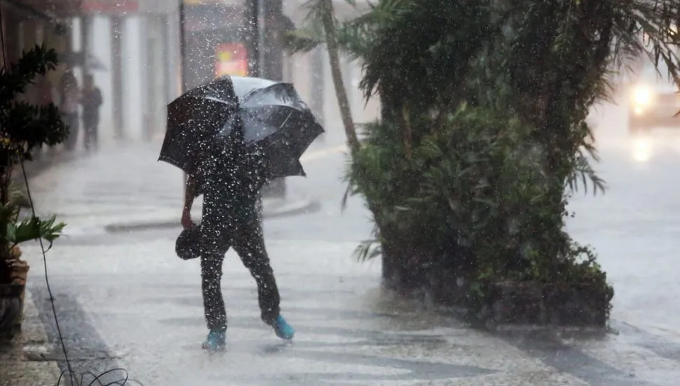 Na imagem, uma pessoa enfrenta chuva forte com guarda-chuva.