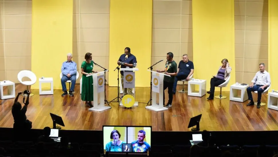 Na imagem, candidatos à prefeitura se enfrentam em um debate, em púlpitos em um palco.