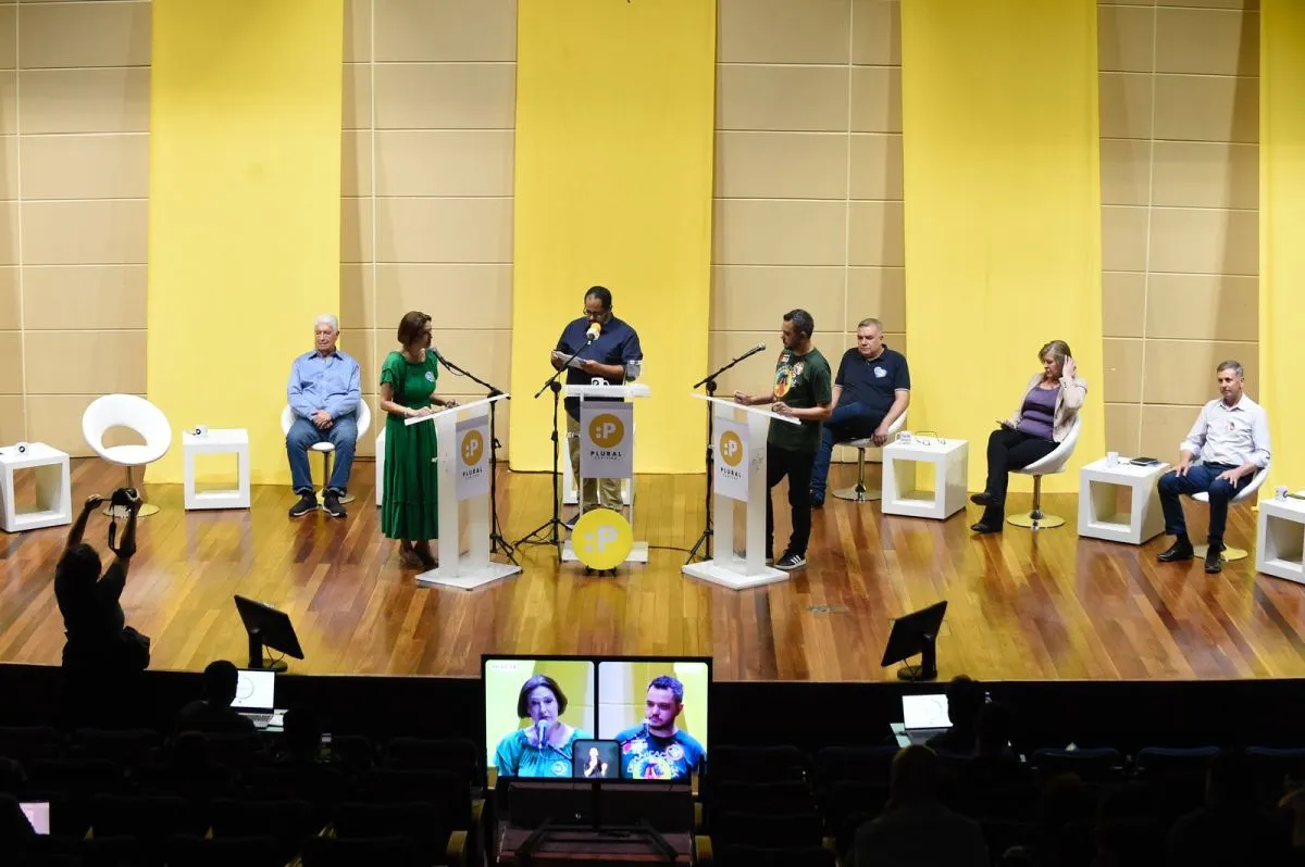 Na imagem, candidatos à prefeitura se enfrentam em um debate, em púlpitos em um palco.